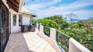Un balcon doté d’un espace repas, offrant une vue sur une végétation luxuriante, et un bâtiment à la façade blanche et au toit végétalisé.