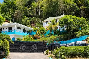 A gated entrance with a blue and white building, surrounded by lush greenery and palm trees.