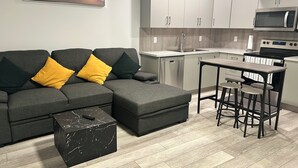 A modern living room with a grey sectional sofa, a black marble coffee table, and a kitchen area featuring a microwave, oven, and bar stools. - New, spacious and lovely two-bedroom basement suite in a peaceful neighbourhood  (Calgary)