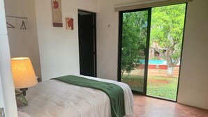 A bedroom with a bed, a bedside table with a lamp, a digital thermometer, and a sliding glass door leading to a garden. - Quinta Yaaxbe (Homún)