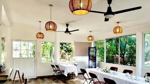 Un espace repas avec une longue table dressée pour un repas, entourée de chaises, et agrémenté de ventilateurs de plafond et de luminaires suspendus.