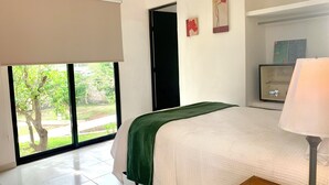 Une chambre d’hôtel avec un lit, une table de chevet, une lampe, une fenêtre donnant sur un espace verdoyant et une porte.