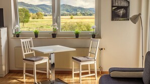 A room with a table and two chairs, a radiator, a window with a view of fields and mountains, and a lamp on the right.