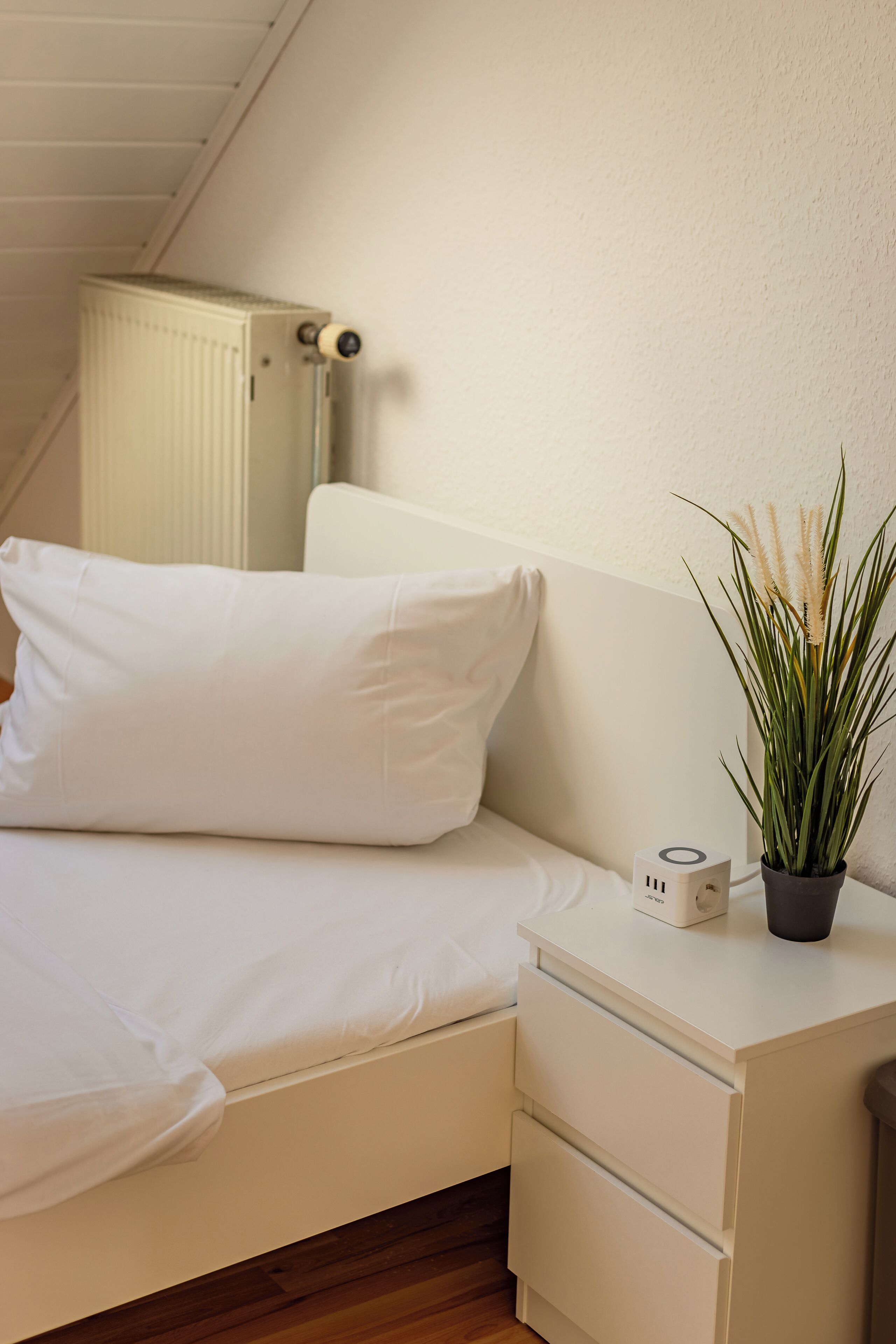 A neatly made bed with white bedding, a bedside table with a small plant, and a radiator against the wall.