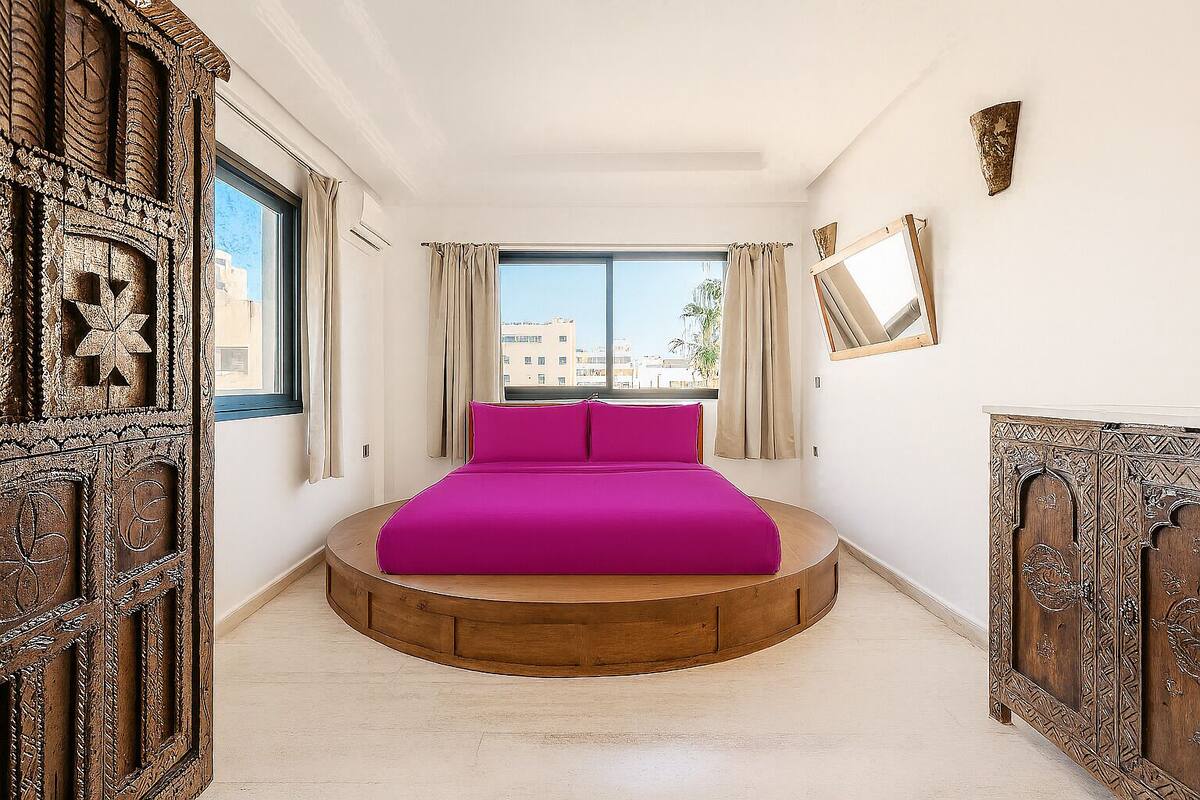 A room with a circular wooden bed platform, a large window with curtains, and a view of buildings outside.