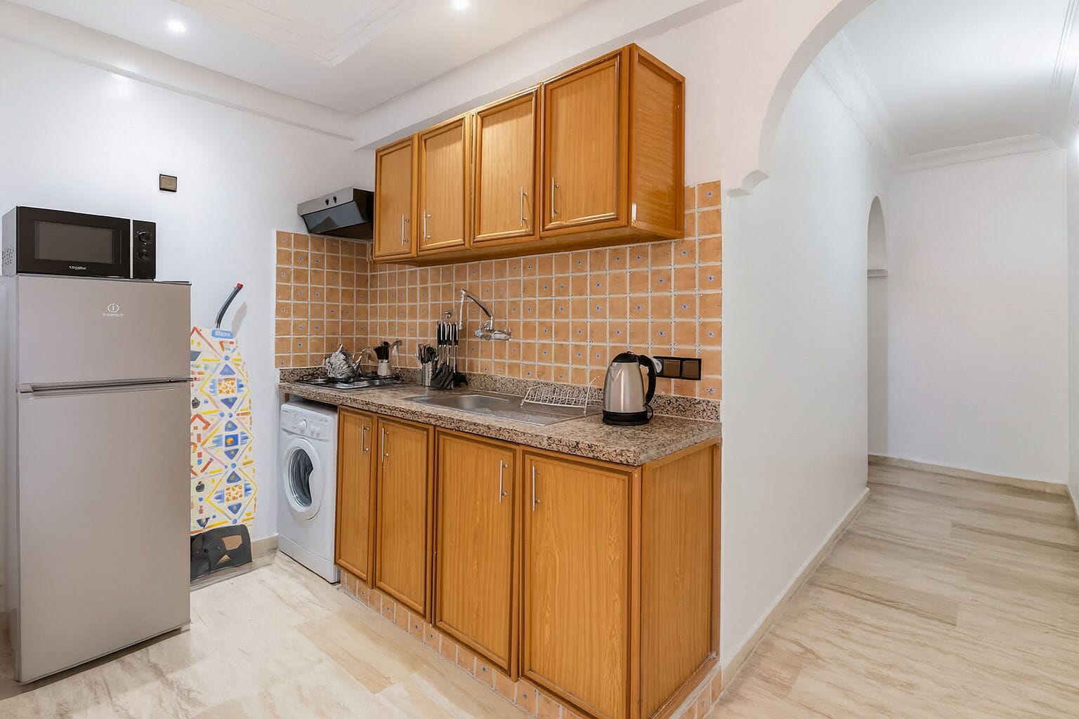A modern kitchen with wooden cabinets, a stainless steel refrigerator, a microwave, a kettle, and a washing machine.