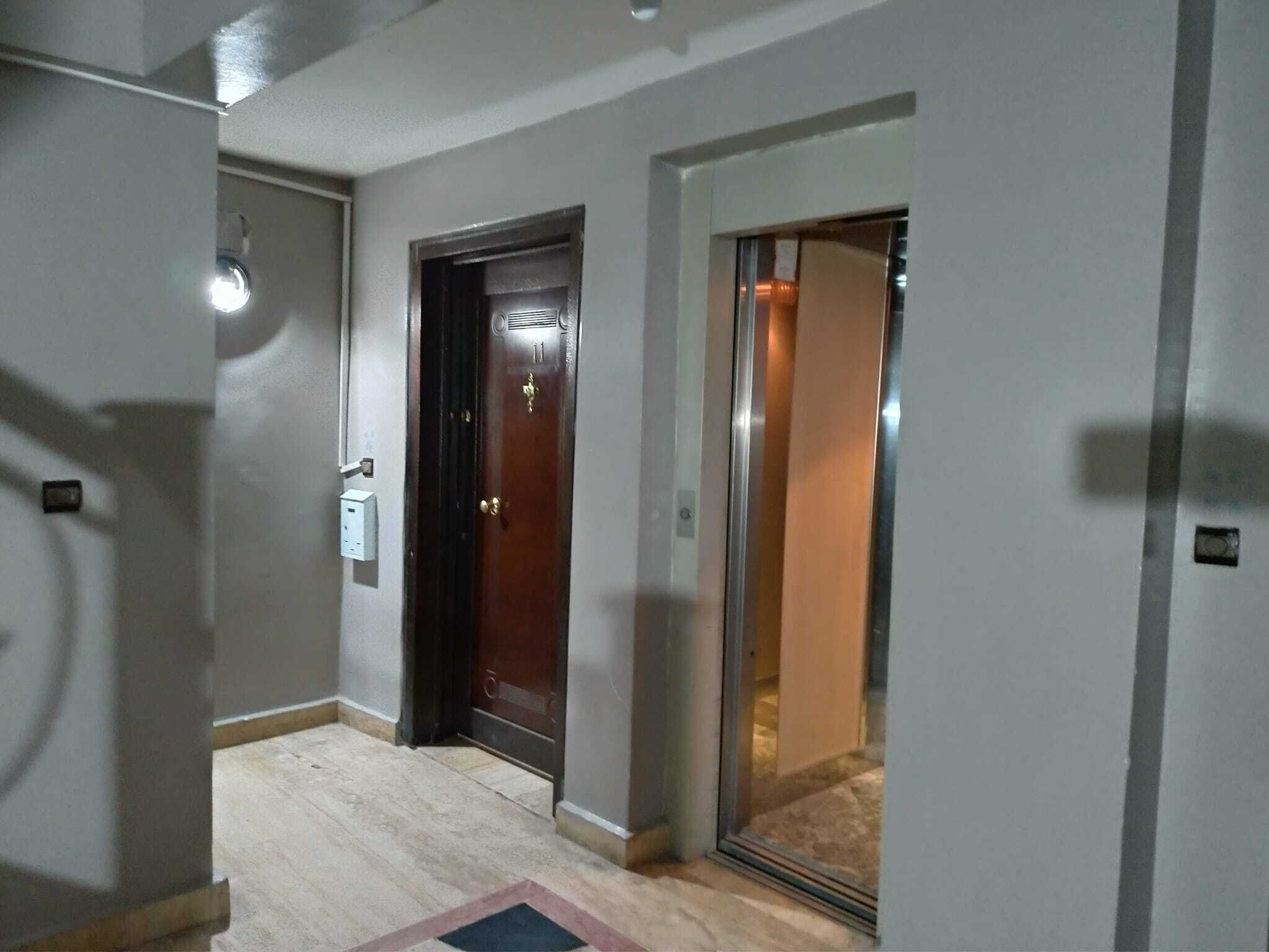 A hallway with a wooden door, a glass elevator, and a light switch on the wall.