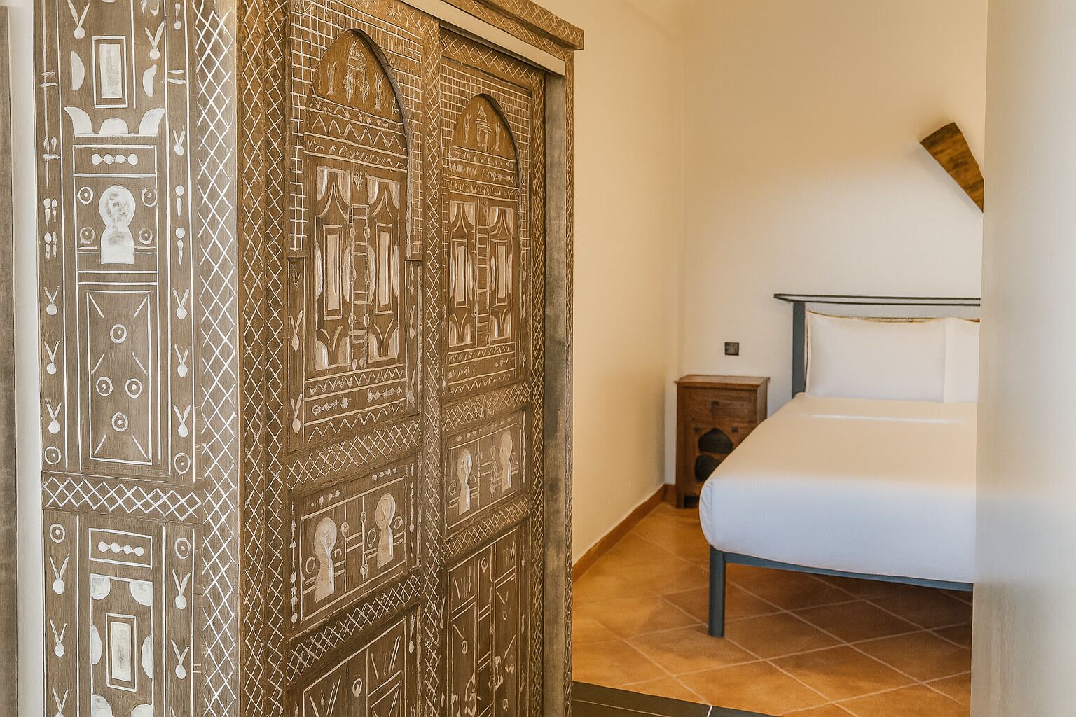 A bedroom with a bed, a wooden wardrobe with intricate designs, and a small wooden nightstand.