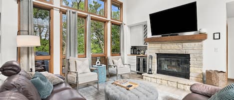 A spacious living room with a stone fireplace, large windows, leather sofas, and a flat-screen TV.