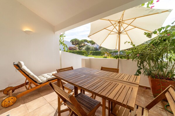 Eine Sonnenliege, ein Holztisch und ein Sonnenschirm auf einer Terrasse mit Blick auf Grünflächen und Hügel.