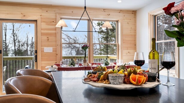 Un interior de madera con una mesa de comedor dispuesta con comida y vino, y vista a los árboles afuera.