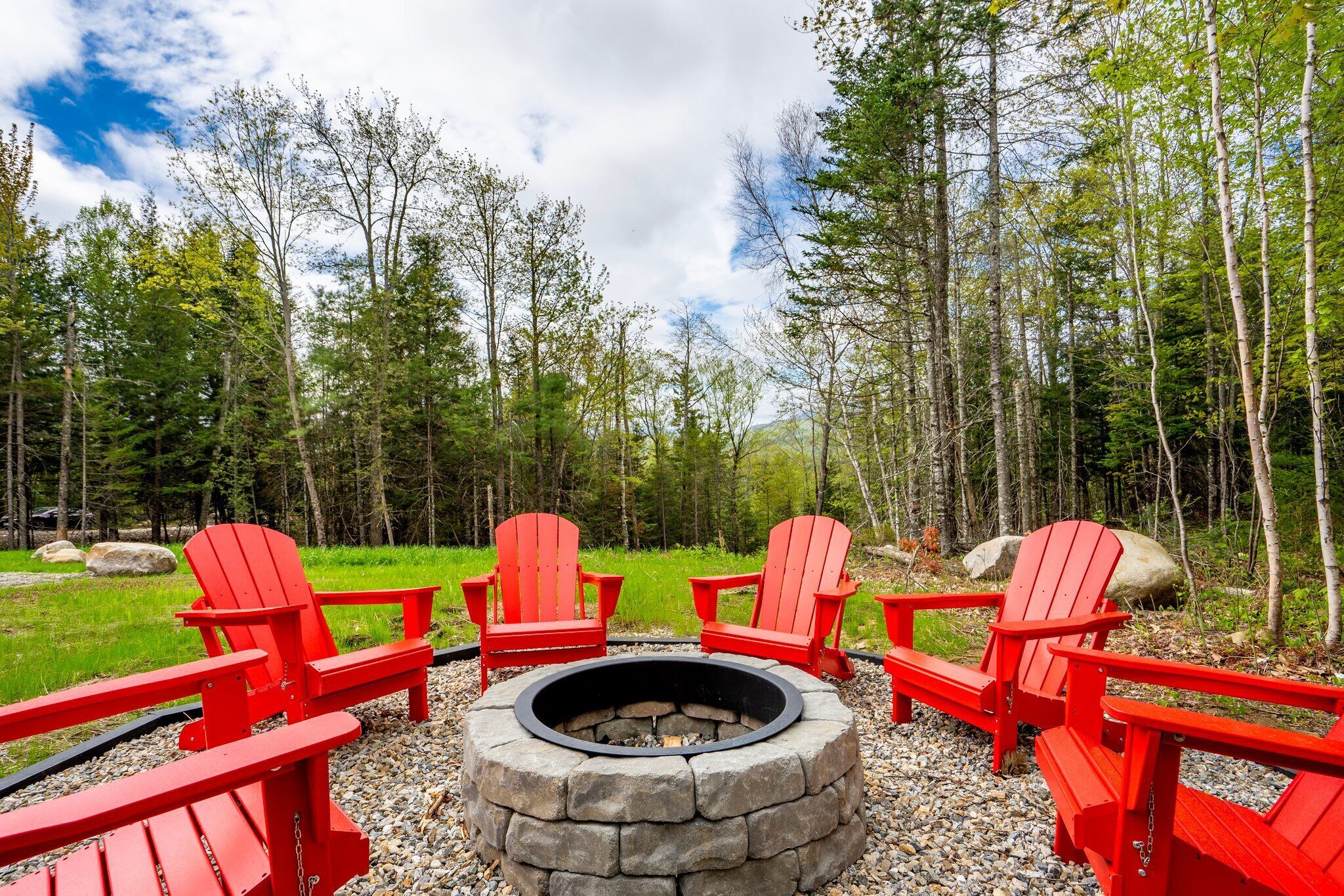 A circular stone fire pit surrounded by four red Adirondack chairs in a forested area.