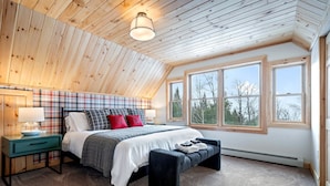 Un dormitorio con techo de madera, cabecera a cuadros, cama con ropa blanca y almohadas rojas, un banco y ventanas amplias con vistas a árboles.