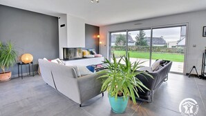 Una sala de estar moderna con un sofá gris, una planta grande en maceta y vistas a un jardín a través de amplios ventanales correderas.