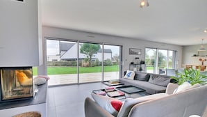 Una sala moderna con chimenea, sofá gris, mesa de centro y amplios ventanales que ofrecen vista al jardín y casas vecinas.