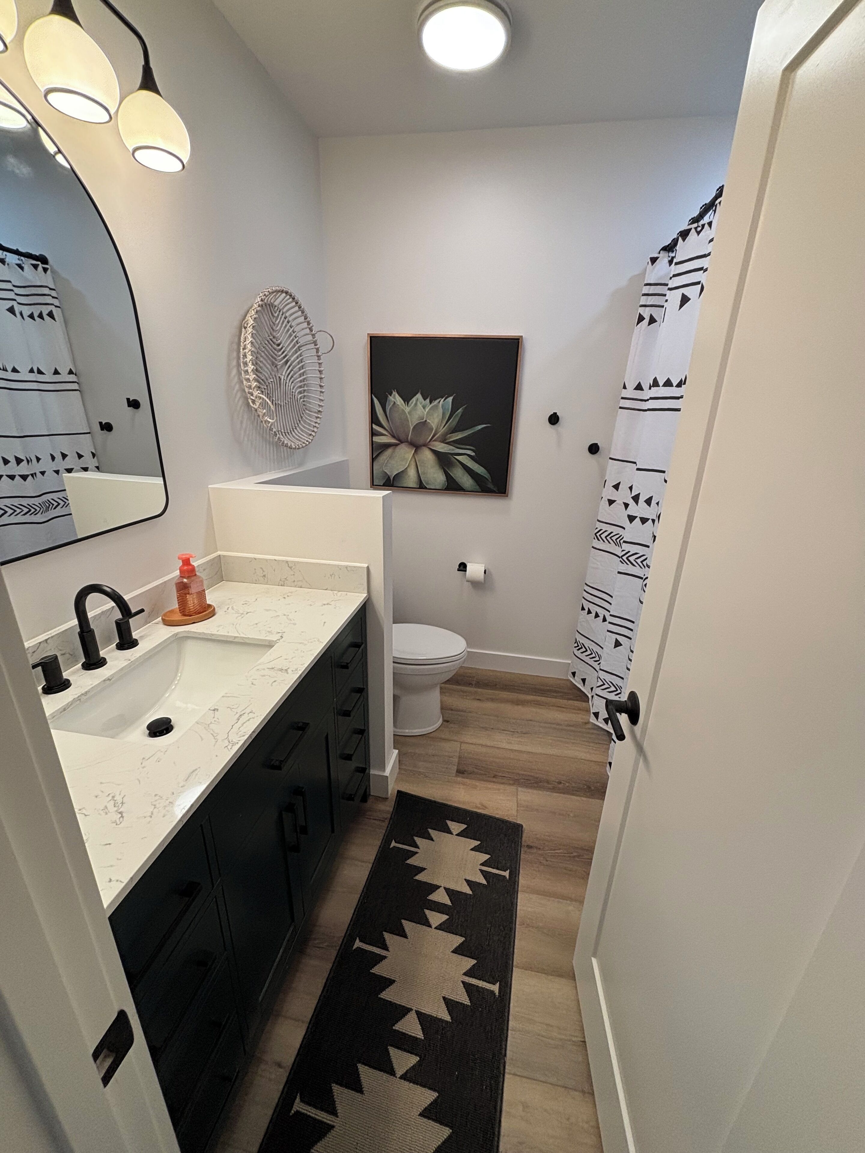 Un baño moderno con un lavamanos blanco, grifo negro y un espejo con tres luces. Se observa un inodoro con tapa blanca y un dispensador de papel higiénico negro. Se ve una cortina de ducha con estampado blanco y negro.