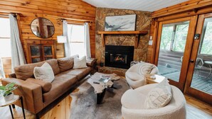 A cozy cabin interior with a stone fireplace, wooden walls, a leather sofa, and a round coffee table. - Romantic Cabin + Hot Tub + Firepit (Ellijay)