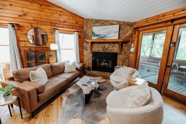 A cozy cabin interior with a stone fireplace, wooden walls, a leather sofa, and a round coffee table. - Romantic Cabin + Hot Tub + Firepit (Ellijay)