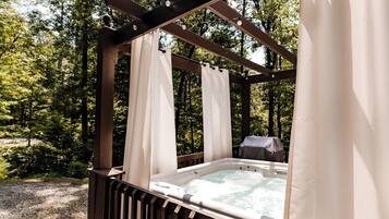A hot tub under a wooden pergola with white curtains, surrounded by a forested area.