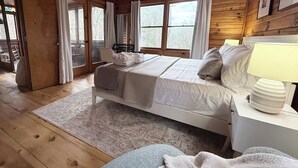 A wooden cabin bedroom with a bed, a bedside table with a lamp, a window with curtains, and a view of trees outside. - Romantic Cabin + Hot Tub + Firepit (Ellijay)