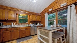 A wooden cabin kitchen with wooden cabinets, a granite countertop, a sink, a dishwasher, and a window with curtains. - Romantic Cabin + Hot Tub + Firepit (Ellijay)