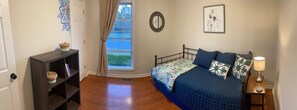 A bedroom with a bed, a nightstand, a lamp, a window with curtains, a mirror, and a wall with a framed picture.