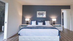 A bedroom with a large bed, two bedside tables with lamps, a ceiling fan, and a door leading to another room. - Captain Dan's Sunset Suite (St. Andrews)