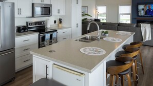 A modern kitchen with white cabinets, stainless steel appliances, and a central island with bar stools. - Captain Dan's Sunset Suite (St. Andrews)