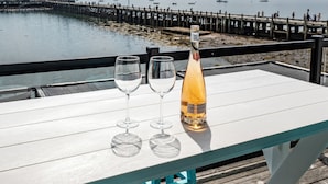 A table with two empty wine glasses and a bottle of champagne on a balcony overlooking a waterfront with a wooden pier. - Captain Dan's Sunset Suite (St. Andrews)