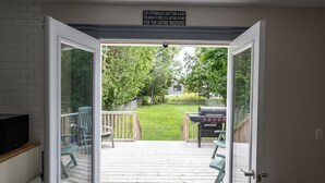 En åben dør fører ud til en træterrasse med grill og stole, et fjernsyn på væggen og et skilt over døren.