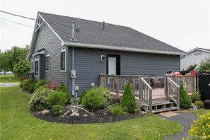 A two-story house with a grey exterior, a front deck with a staircase, and a landscaped garden with various shrubs and a small tree. - Captain Dan's Cozy on Queen (St. Andrews)