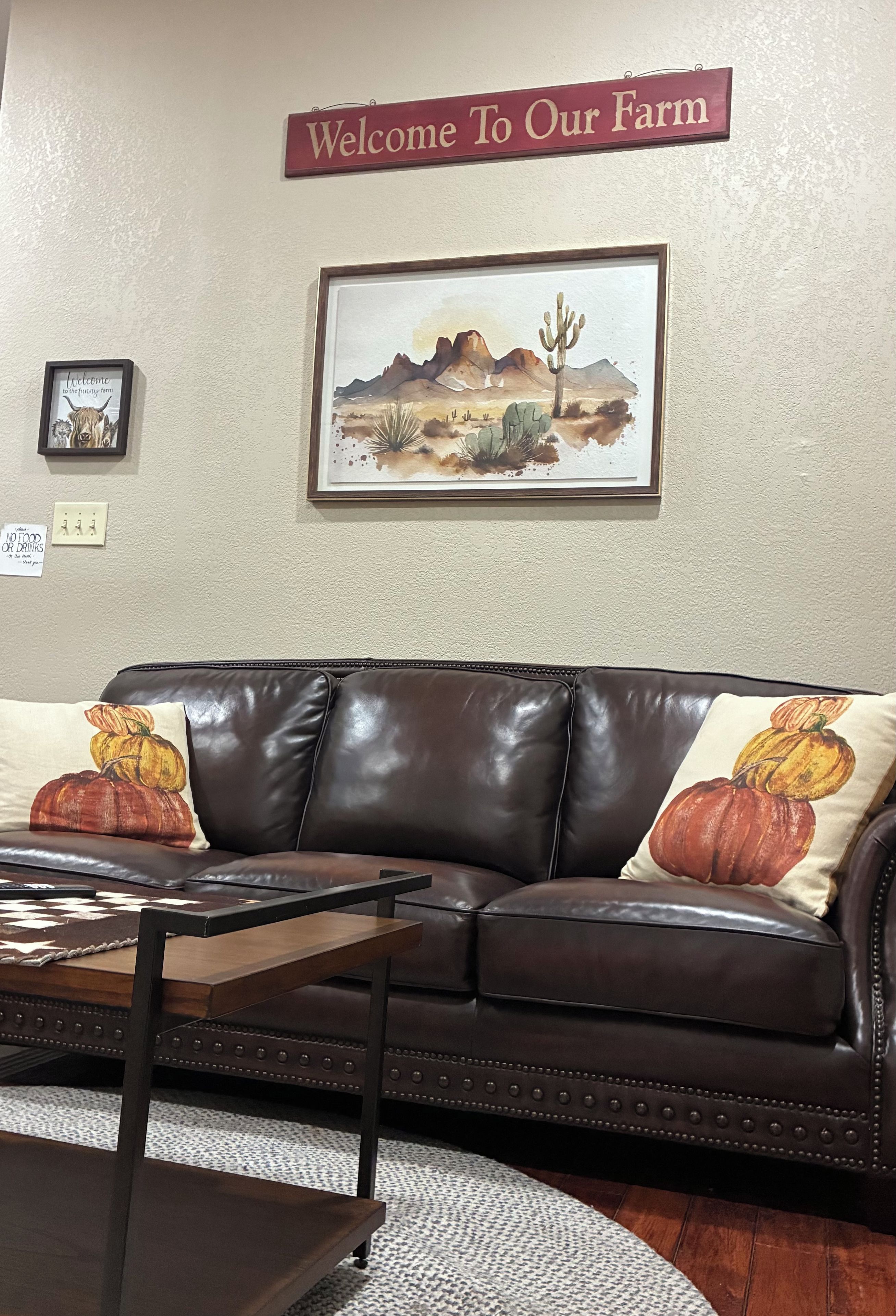 Una sala de estar con un sofá de cuero marrón, dos cojines con diseños de calabaza, un cuadro de paisaje enmarcado y un cartel de bienvenida sobre el sofá.