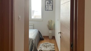 Un couloir mène à une chambre qui contient un lit, une fenêtre, un radiateur et une œuvre d’art encadrée accrochée au mur.
