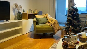 Un salon moderne avec un fauteuil vert, un téléviseur posé sur un meuble, un sapin de Noël et une vue sur la ville.