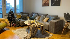 Un salon moderne avec un canapé gris, une petite table ronde décorée de bougies, un sapin de Noël et une vue sur la ville, visible à travers de grandes fenêtres.