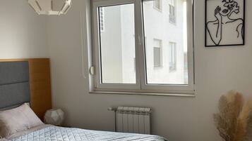 Une chambre avec un grand lit, un radiateur, une fenêtre avec des stores, un luminaire suspendu et une œuvre d’art murale.