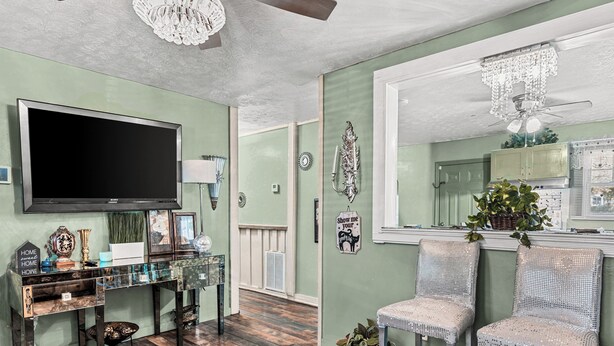 A room with a large flat-screen TV, a glass display table, two upholstered chairs, a ceiling fan, and a decorative mirror.