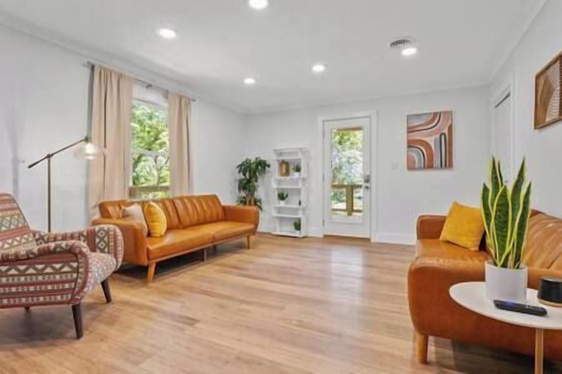 Un salón amplio con suelo de madera, un sofá de cuero marrón, un sillón estampado y un estante blanco.
