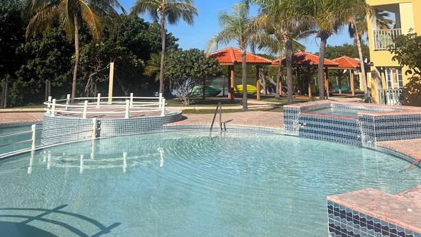 A clear blue sky, a swimming pool with tiled edges, palm trees, and a yellow building with a red roof. - Sol y Mar - Oceanfront Condo! (Cabo Rojo)
