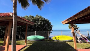 A clear view of a beachside area with a palm tree, a fenced area with colorful inflatable rafts, and a wooden structure with a red roof. - Sol y Mar - Oceanfront Condo! (Cabo Rojo)