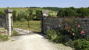 A gated entrance with a stone wall and a metal gate, leading to a green field and trees. - Le Cellier aux Moines (COLOMBE LE SEC)
