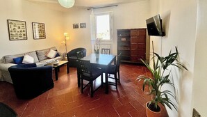 Un salon comprenant un canapé, une table à manger, des chaises, un téléviseur fixé au mur et une plante en pot.