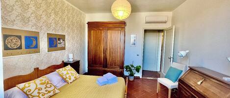 A bedroom with a bed, a wooden wardrobe, a desk, a chair, and a hanging light fixture.