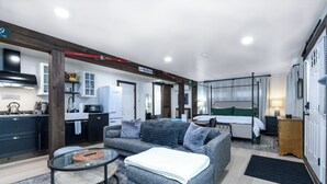 A modern hotel room with a grey sofa, a glass coffee table, a bed with green bedding, a small wooden nightstand, and a kitchen area visible through an open door.