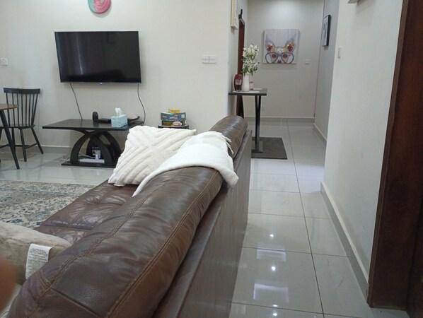 A living room with a brown leather sofa, a flat-screen TV mounted on the wall, a small table with a vase of flowers, and a painting on the wall.