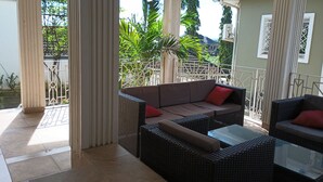 A covered outdoor seating area with wicker furniture and a glass table, surrounded by white railings and greenery.