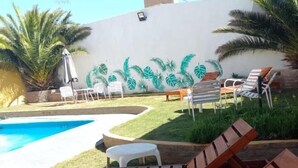 A poolside area with wooden loungers, a white wall with a tropical leaf mural, and a small white table. - Buenavista Hotel (La Falda)