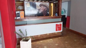 A reception desk with a logo, a framed picture, and a potted plant. - Buenavista Hotel (La Falda)