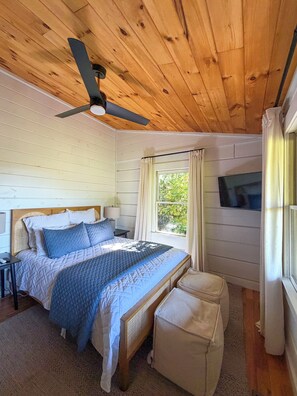 A bedroom with a bed, a ceiling fan, a TV, a window with curtains, and a small table.