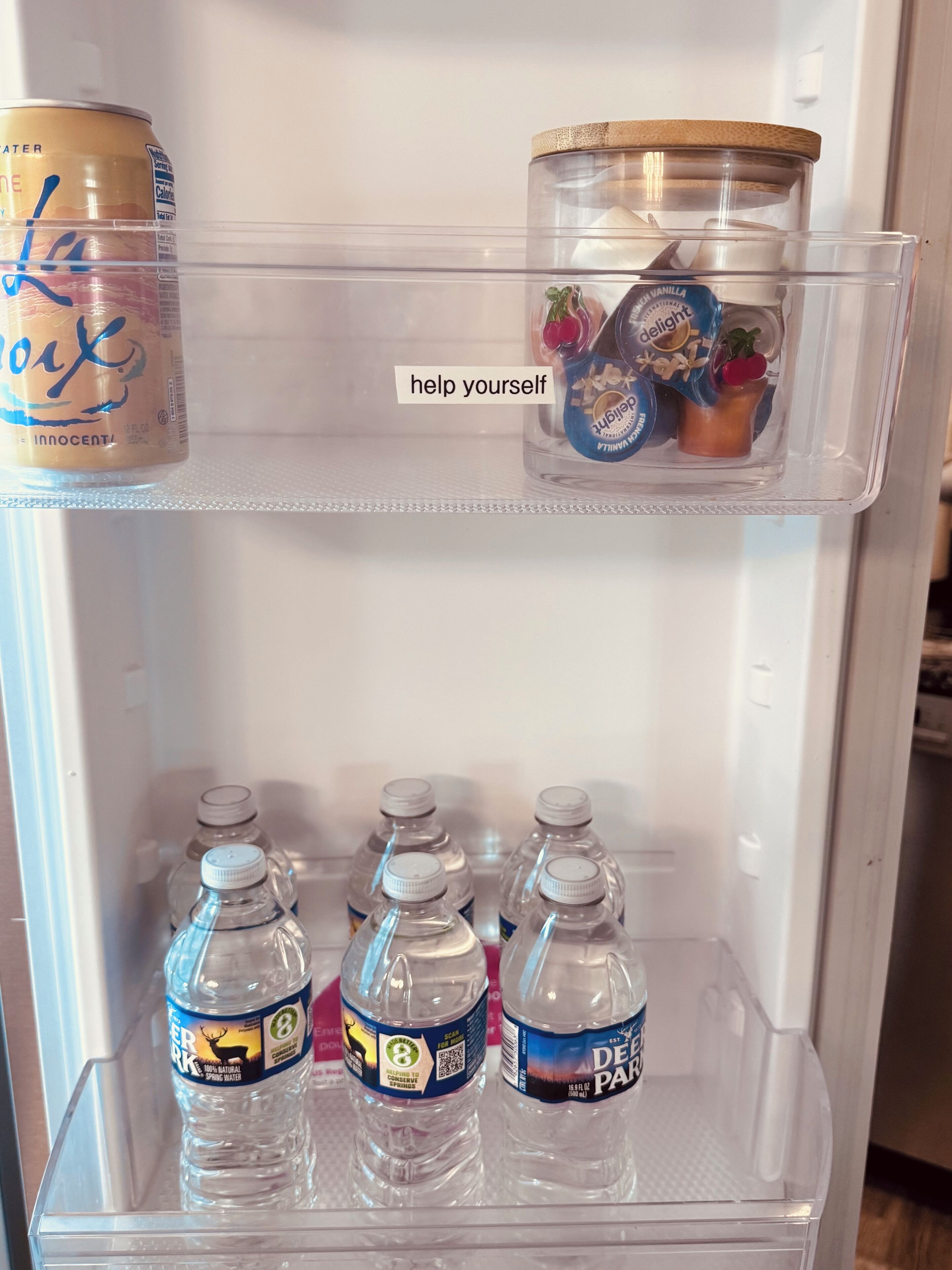 Un refrigerador con un estante transparente que contiene una lata, un frasco con adornos y tres botellas de agua.